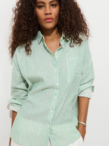 A woman with curly hair wears the Jo Shirt In Double Gauze Stripe—mint green and white stripes, relaxed fit, sleeves rolled up—standing against a plain white background.