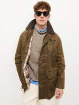 A person wearing sunglasses, the Regent Jacket in Waxed Cotton with a plaid lining, a gray knit sweater, a blue collared shirt, and light pants stands against a plain white background.
