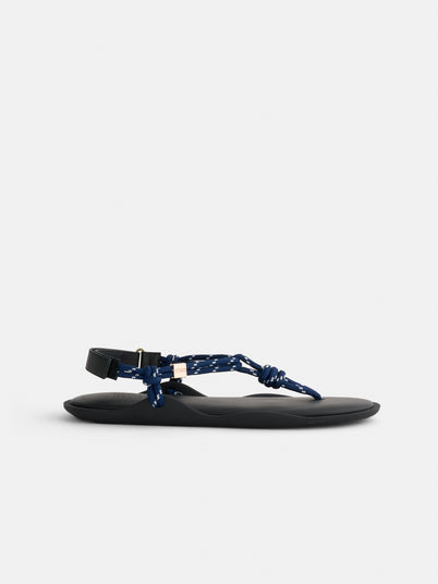 Flo Sandals feature a flat black recycled rubber sole and blue rope straps with white speckles and knotted detailing, inspired by Tuscany sandals, and are secured at the ankle with a black buckle.