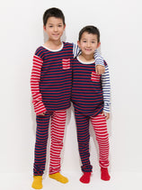 Two young boys stand side by side, smiling with arms around each other’s shoulders. They wear matching Mini Sleep Sets—100% cotton pajamas with red, white, and blue stripes—and colorful socks, posing against a plain white background.