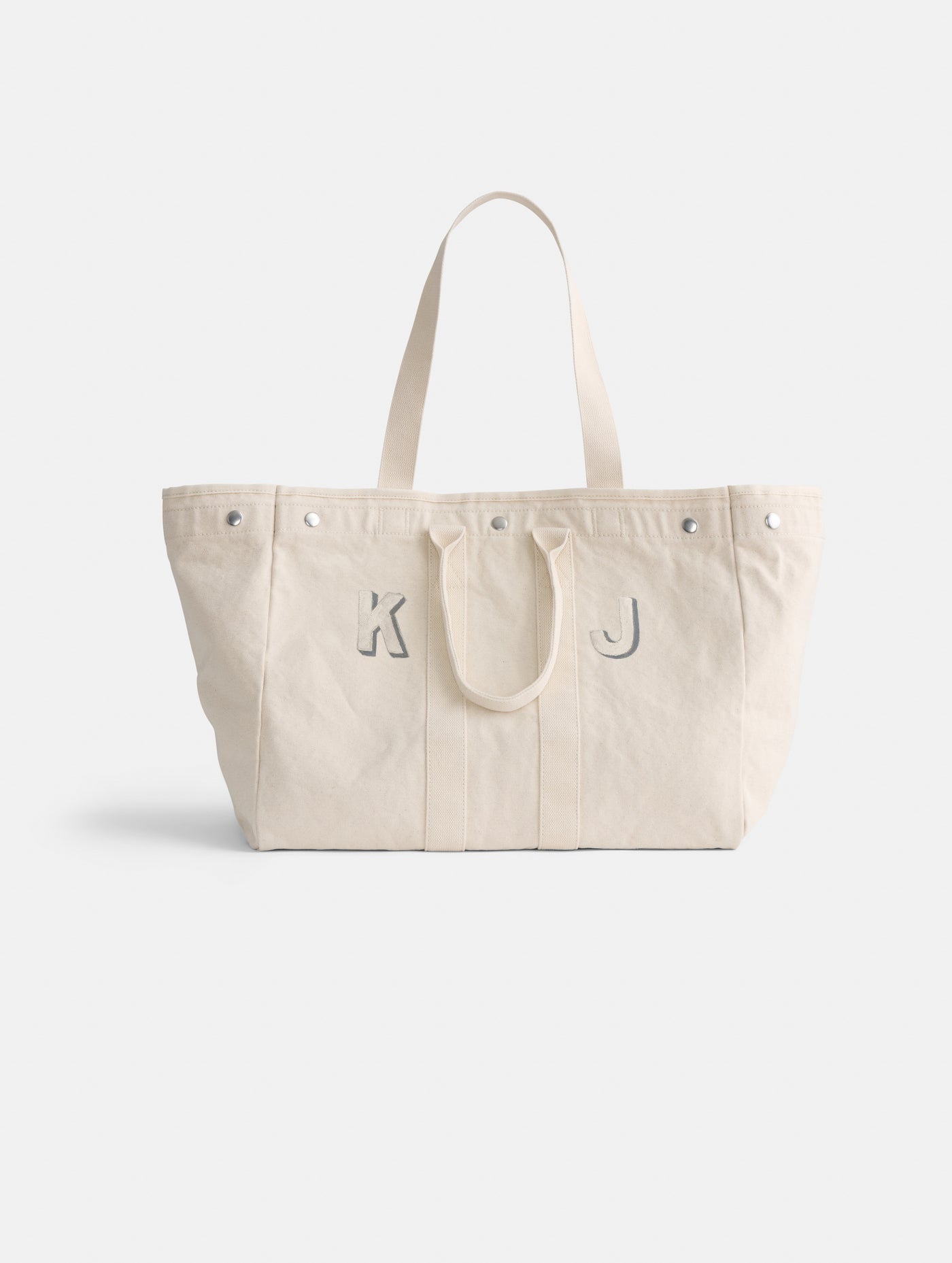 The Painted Perfect Weekend Tote is a spacious off-white canvas bag with two handles, silver rivets along the top edge, and light gray 