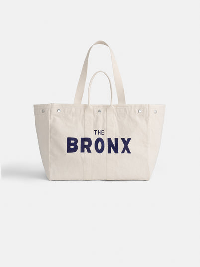 The Bronx Painted Perfect Weekend Tote is a large, custom-made off-white bag with two handles and bold, dark blue “THE BRONX” hand-painted on the front.