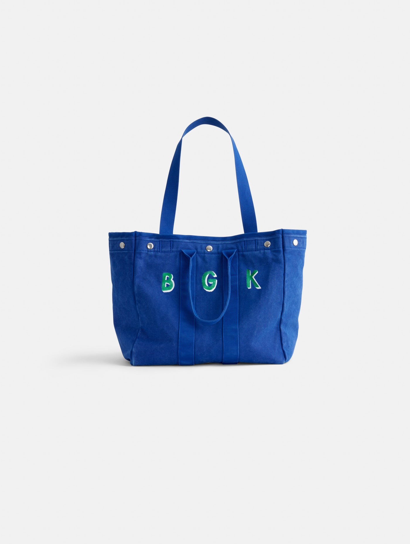 The Perfect Weekday Tote is a blue cotton bag with two handles and snap closures. Green and white embroidered letters B G K add a personal touch to this monogrammable tote, shown on a plain white background.