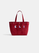 The Painted Perfect Weekday Tote is a large red canvas bag with two handles and “E L I” in white and blue on the front, displayed against a plain white background.