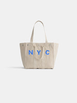 The Perfect Weekday Tote is a beige cotton bag with two handles and bold blue NYC lettering on the front, shown against a white background. Ideal for anyone looking for a stylish and customizable everyday tote.