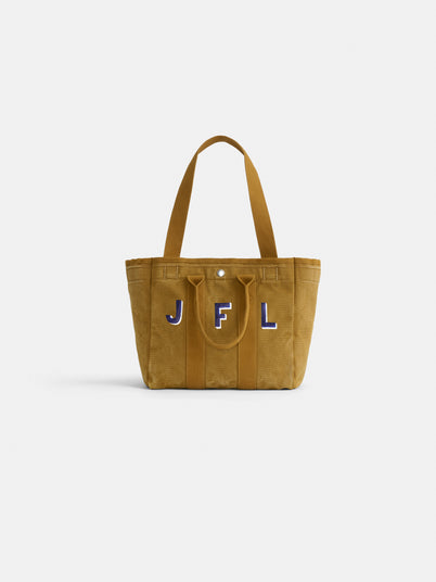 The Perfect Daily Tote, a tan suede bag with two handles and blue-and-white JFL embroidery on the front, combines classic style and monogram versatility. Shown on a plain white background.