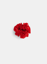 A red carnation with ruffled petals sits on a plain white background beside the Frisée Hair Scrunchie In Linen—a sustainable, zero waste hair accessory that blends beauty with eco-conscious style.