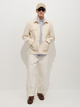 A person wearing the Garment Dyed Work Jacket In Recycled Denim, a striped blue-and-white shirt, white pants, a beige baseball cap, dark sunglasses, and brown shoes stands against a plain white background with hands in jacket pockets.