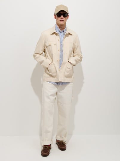 A person wearing the Garment Dyed Work Jacket In Recycled Denim, a striped blue-and-white shirt, white pants, a beige baseball cap, dark sunglasses, and brown shoes stands against a plain white background with hands in jacket pockets.
