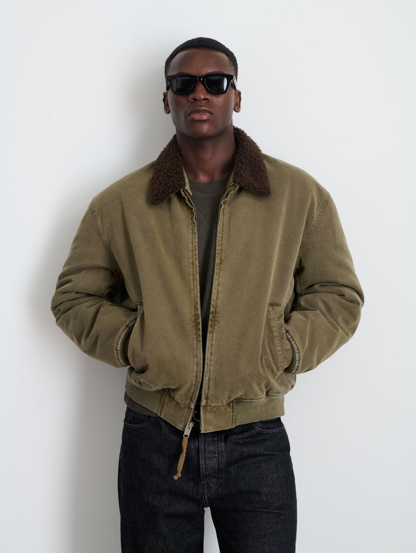 A man wearing dark sunglasses, black jeans, and the Flight Jacket In Canvas with an olive green sherpa collar and quilted lining stands against a plain white background, hands in his pockets.