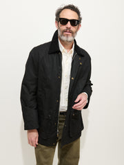 A man with a beard and mustache, wearing black sunglasses, the Regent Jacket in Waxed Cotton, a white shirt, and olive green pants stands against a plain white background.