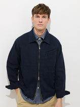 A young man with short brown hair stands against a white background, wearing the navy Andre Zip Jacket In Linen Twill over a blue button-up shirt and beige pants. He looks directly at the camera with a neutral expression.