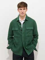 A young man with short brown hair wears the Riviera Work Jacket in Linen over a white collared shirt and black pants, standing against a plain white background with his hands in his pockets.