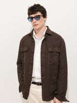 A man wearing the Riviera Work Jacket in Linen over a white button-up shirt, light-colored pants, and blue sunglasses stands against a plain white background with one hand in his pocket.