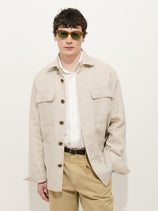 A man wearing green sunglasses, the Riviera Work Jacket in flax linen with faux horn buttons, a white shirt, and khaki pants stands against a plain white background with one hand in his pocket.