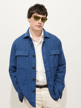 A man wearing green sunglasses and the Riviera Work Jacket In Linen over a white shirt with light pants stands against a plain white background, hands in his pockets.