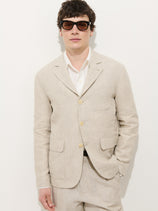 A man wearing dark sunglasses, a white shirt, and the Mercer Linen Jacket in Flax stands against a plain white background with one hand in his pocket.