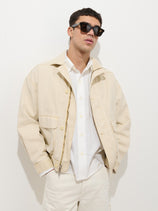 A man wearing black sunglasses, the Giorgio Bomber Jacket In Canvas in light beige over a white button-up shirt, and matching beige pants stands against a plain white background with his hands in his pockets.