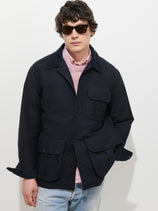 A person wearing sunglasses, the Clifton Jacket In Moleskin over a pink shirt, and light blue jeans stands against a plain white background with hands in their pockets.
