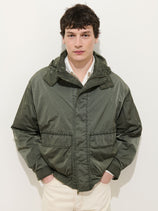 A young man with short dark hair stands against a white background, wearing the David Windbreaker in crinkle nylon with a removable hood, paired with a white collared shirt and cream pants. His hands rest in the windbreaker's pockets.