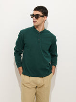 A man wearing the Alan Henley In Heavyweight Cotton in dark green, beige pants, and black sunglasses stands against a plain white background with his hands in his pockets.