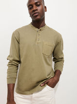 A man in the Alan Henley In Heavyweight Cotton, featuring an olive green vintage wash and chest pocket, pairs it with light-colored pants and stands against a plain white background with one hand in his pocket.