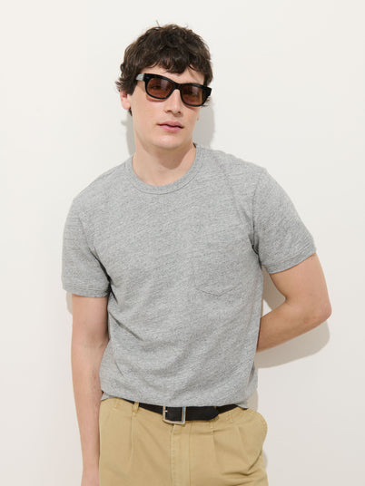 A man wearing dark sunglasses, beige pants, and the Standard Pocket Tee in Slub Cotton in light gray stands against a plain white background with one hand behind his back.