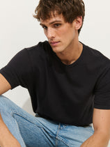 A young man with short brown hair wears The Standard T-Shirt In Slub Cotton and light blue jeans, sitting with one knee up and gazing slightly to the side against a plain white background.
