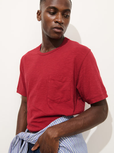 A man wearing the Standard Pocket Tee in Slub Cotton, featuring a chest pocket and a vibrant red hue, looks down and to the side. He has a blue and white striped shirt tied around his waist and stands against a plain white background. Made in LA, this breathable t-shirt combines effortless comfort with laid-back style.