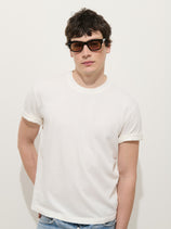 A young man wearing dark sunglasses and the Crosby Tee in Midweight Jersey stands against a plain white background in blue jeans, looking slightly to the side with a relaxed expression.