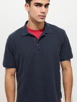 A man wears the Garment Dyed Polo In Pique Cotton in navy blue over a red T-shirt, standing against a plain white background and looking slightly to the side with a neutral expression.