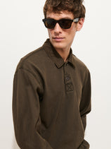 A man wearing dark sunglasses and a dark olive Garment Dyed Rugby Shirt In Cotton Jersey with a boxy fit looks to the side against a plain white background.