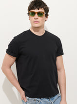 A person wearing green sunglasses, the Crosby Tee In Midweight Jersey—an easy fit black 100% cotton rolled-sleeve tee—and light blue jeans stands against a plain white background with one hand in their pocket.