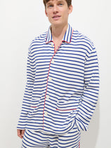 A man wearing The Striped Waffle Sleeping Shirt—a blue and white striped 100% cotton pajama top with red trim—stands against a plain white background, one hand in his pocket and a slight smile on his face.