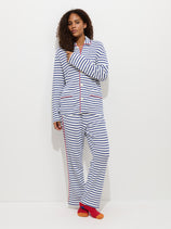 A woman with curly hair stands against a white background wearing The Striped Waffle Sleeping Shirt, featuring blue and white stripes, red buttons, and accents. She completes her cozy look with mismatched orange and red socks.