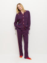 A person stands against a white wall wearing The Striped Waffle Pant—blue and red striped 100% cotton pajamas with white buttons—and orange-accented socks, looking to the side with hands in their pockets.