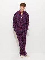 A person stands against a white background, wearing The Striped Waffle Pant in blue and red with white buttons. Their hands are in their pockets, and they are also wearing white slippers.