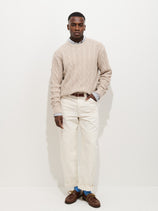 A man stands against a white background wearing the Aaron Cable Sweater in Cotton, styled over a collared shirt with white pants, a brown belt, and brown loafers. With his left hand in his pocket, he gazes confidently at the camera.