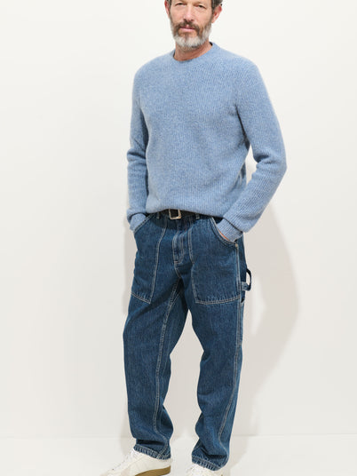 A man with gray hair and a beard wears a light blue sweater, the Painter Pant in Vintage Wash Denim, a black belt, and white sneakers, standing against a plain white background with his hands in his pockets.