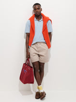 A man stands against a white wall wearing a light blue polo, Pull On Short In Cotton Herringbone with drawstring waist, brown boat shoes, yellow socks, an orange sweater draped over his shoulders, holding a red tote bag and looking at the camera.