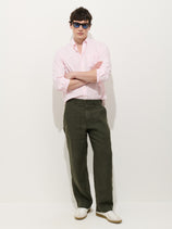 A person stands against a white background wearing a light pink button-down shirt, Pull On Field Pant In Linen in olive green, white sneakers, and blue sunglasses, with arms crossed and a relaxed expression.
