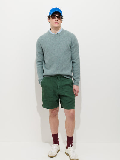 A person stands against a plain white background wearing a blue cap, sunglasses, a light blue sweater over a collared shirt, Garment Dyed Pull On Field Short In Linen, maroon socks, and white sneakers.