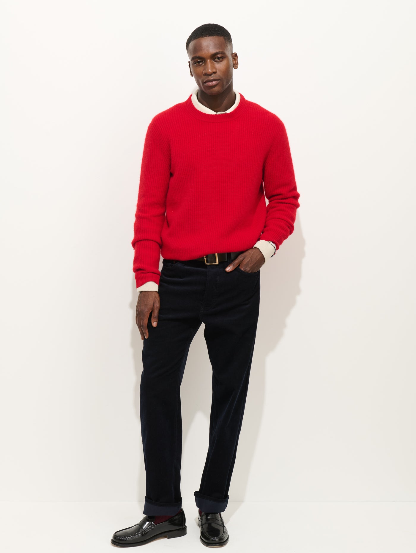 A man with short hair poses against a white background wearing the AM Original 5 Pocket Cord in a straight tapered fit, paired with a red sweater over a white collared shirt and black loafers, one hand casually in his pocket.