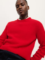 A person wears the Jordan Sweater In Washed Cashmere, a vibrant red and ultra-soft piece, paired with dark pants and poses confidently against a plain white background.