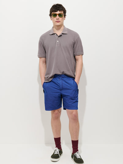 A man wears sunglasses, a gray polo shirt, Camino Short in Crinkly Nylon (vintage-inspired bright blue), maroon socks, and black sneakers, standing against a plain white background with his hands in his pockets.
