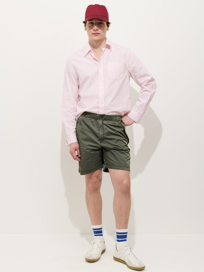 Against a white background, a person wears a red cap, light pink button-up shirt, Camino Short In Crinkly Nylon, white sneakers, and white socks with blue stripes. One hand is in their shorts pocket as they look at the camera.