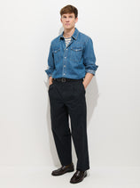 A man stands against a white background wearing black Georges Pull On Pant in lightweight cotton with a belted waist, paired with a blue denim shirt over a black-and-white striped t-shirt and dark brown dress shoes, hands in his pockets.