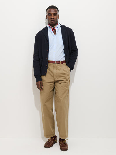 A man stands against a plain white background wearing the Georges Pull On Pant In Lightweight Cotton, a navy cardigan, light blue button-up shirt, red patterned neckerchief, brown leather belt, and brown lace-up shoes. One hand is in his pocket.