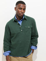 A man with short hair and a neutral expression stands against a white background, wearing the Carter Popover In Cotton Linen—a green men's shirt with shell buttons—over a blue collared shirt and light brown pants.