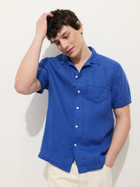 A young man stands against a white background, wearing the Camp Shirt In Linen with light-colored pants, one hand in his pocket and the other touching his head.